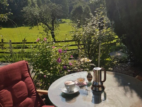 Garten.jpg Gemütlicher Gartenstuhl und Tisch mit Kaffeekanne und Tassen. Blühender Garten, üppige Bäume und Hügel.