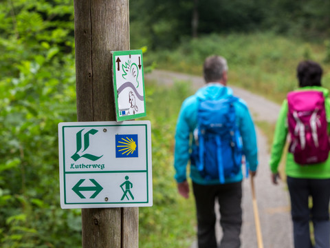Schild, auf dem Lutherweg steht, dahinter verschwommen ein Paar in WanderkleidungSign saying Lutherweg, behind it a blurred couple in hiking clothes