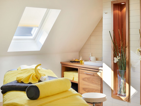Wellnesspark Elbresidenz Heller Massageraum mit gelber Liege, Handtüchern und Orchidee, Dachfenster, Holzschrank mit Waschbecken, entspannte Atmosphäre.