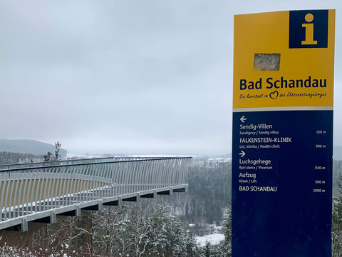 Skywalk Bad Schandau im Winter Gelbes Informationsschild mit der Aufschrift "Bad Schandau" vor einer verschneiten Landschaft und einer modernen Brücke im Hintergrund.