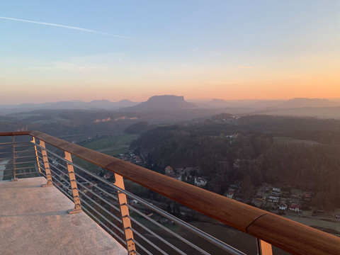 Aussicht von einem Balkon mit Holzgeländer auf eine hügelige Landschaft im Sonnenuntergang, der Himmel ist blau-orange gefärbt.