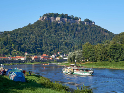 Ein Fluss mit einem Dampfschiff im Vordergrund, dahinter ein bewaldeter Hügel mit der Festung Königstein; Zelte am Ufer, klare, sonnige Atmosphäre.A river with a steamboat in the foreground, behind it a wooded hill with the Königstein fortress; tents on the bank, clear, sunny atmosphere.Řeka s parníkem v popředí, za ní zalesněný kopec s pevností Königstein, stany na břehu, jasná, slunečná atmosféra.Rzeka z parowcem na pierwszym planie, za nią zalesione wzgórze z twierdzą Königstein; namioty na brzegu, czysta, słoneczna atmosfera.Een rivier met een stoomboot op de voorgrond, daarachter een beboste heuvel met het fort Königstein; tenten aan de oever, heldere, zonnige sfeer.Un fiume con un battello a vapore in primo piano, alle spalle una collina boscosa con la fortezza di Königstein; tende sulla riva, atmosfera limpida e soleggiata.