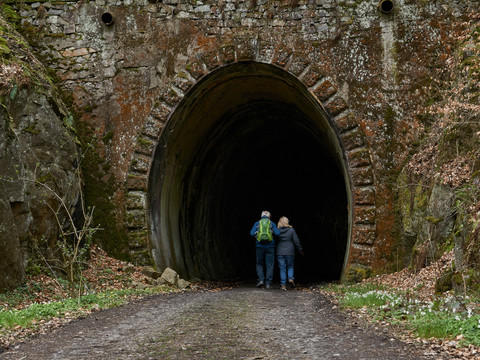 Former tunnel of the Schwarzbach Valley Railway Zwei Personen gehen in einen dunklen, moosbewachsenen Tunnel aus Stein; die Umgebung ist bewaldet und herbstlich.