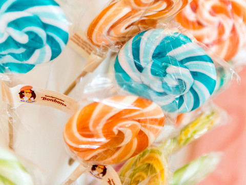 Lollis aus der Bonbonmanufaktur Bunte Lollipops in Blau-Weiß und Orange-Weiß, in Zellophan verpackt, mit Etikett "handgemachte Bonbons" auf einem Holzstiel.