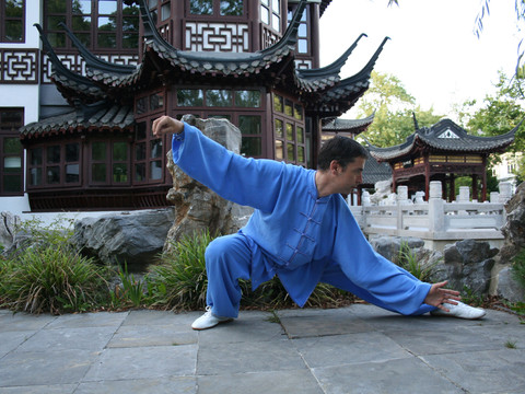 Taijiquan Saksisch Zwitserland Ein Mann in blauer Kampfkunstkleidung führt eine Tai-Chi-Position in einem traditionellen chinesischen Garten mit Pavillon im Hintergrund aus.