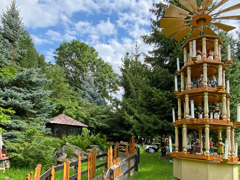 Aussenpyramide.jpg Große, mehrstöckige Weihnachtspyramide mit Figuren und Kerzen in einem grünen Garten mit Tannenbäumen und blauem Himmel im Hintergrund.