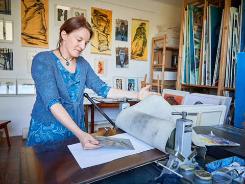 Künstlerin Anne Kern Eine Frau in blauer Kleidung arbeitet in einem Atelier an einer Druckpresse, umgeben von Kunstwerken und Malutensilien.A woman in blue clothing works at a printing press in a studio, surrounded by works of art and painting utensils.Žena v modrém oblečení pracuje v ateliéru u tiskařského lisu, obklopena uměleckými díly a malířskými potřebami.Kobieta ubrana na niebiesko pracuje przy prasie drukarskiej w pracowni, otoczona dziełami sztuki i przyborami malarskimi.Een vrouw gekleed in blauw werkt aan een drukpers in een studio, omringd door kunstwerken en schildergerei.Una donna vestita di blu lavora a una macchina da stampa in uno studio, circondata da opere d'arte e utensili da pittura.
