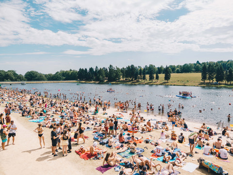 Menschen genießen einen sonnigen Tag am belebten Strand eines Sees mit bewaldetem Ufer.People enjoying a sunny day on the lively beach of a lake with a wooded shore.Lidé si užívají slunečného dne na živé pláži jezera se zalesněným břehem.Ludzie cieszący się słonecznym dniem na tętniącej życiem plaży jeziora z zalesionym brzegiem.Mensen genieten van een zonnige dag op het levendige strand van een meer met een beboste oever.Persone che si godono una giornata di sole sulla vivace spiaggia di un lago con una riva boscosa.