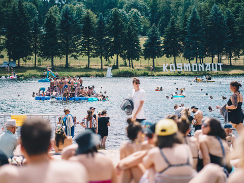 Menschen genießen eine Musikveranstaltung am See unter Bäumen, manche im Wasser, andere am Ufer.People enjoy a music event by the lake under the trees, some in the water, others on the shore.Lidé si užívají hudební akci u jezera pod stromy, někteří ve vodě, jiní na břehu.Ludzie cieszą się imprezą muzyczną nad jeziorem pod drzewami, niektórzy w wodzie, inni na brzegu.Mensen genieten van een muziekevenement bij het meer onder de bomen, sommigen in het water, anderen op de oever.La gente si gode un evento musicale in riva al lago sotto gli alberi, alcuni in acqua, altri sulla riva.
