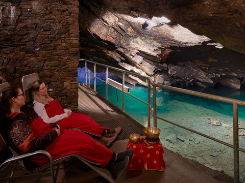 Klangschalenzeremonie im Besucherbergwerk Zwei Personen in roten Decken entspannen auf Liegestühlen in einer Höhle mit türkisfarbenem Wasser, neben ihnen stehen Klangschalen auf einem Tisch.