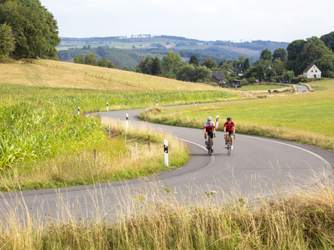Rennradfahren im Bergischen Zwei Radfahrer in sportlicher Kleidung fahren eine kurvige Straße durch eine hügelige Landschaft.