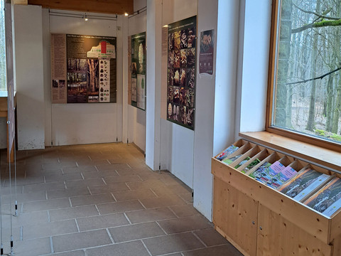 Infomaterial im Erdgeschoss der Infostelle Eishaus Ein Ausstellungsraum mit Informationstafeln über Wälder, einem Holztisch mit Broschüren und einem großen Fenster mit Blick auf den Wald.An exhibition room with information boards about forests, a wooden table with brochures and a large window with a view of the forest.Výstavní místnost s informačními tabulemi o lesích, dřevěným stolem s brožurami a velkým oknem s výhledem na les.Sala wystawowa z tablicami informacyjnymi o lasach, drewnianym stołem z broszurami i dużym oknem z widokiem na las.Een tentoonstellingsruimte met informatieborden over bossen, een houten tafel met brochures en een groot raam met uitzicht op het bos.Una sala espositiva con pannelli informativi sulle foreste, un tavolo di legno con opuscoli e una grande finestra con vista sulla foresta.
