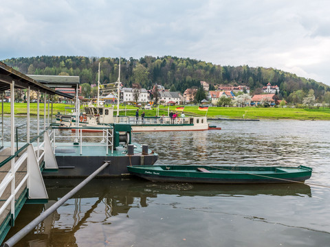 Fähre Kurort Rathen Fähre auf einem Fluss mit grüner Landschaft und Häusern im Hintergrund, bewölkter Himmel, ruhige Atmosphäre.Ferry on a river with green landscape and houses in the background, cloudy sky, calm atmosphere.Přívoz na řece se zelenou krajinou a domy v pozadí, zatažená obloha, klidná atmosféra.Prom na rzece z zielonym krajobrazem i domami w tle, zachmurzone niebo, spokojna atmosfera.Veerboot op een rivier met groen landschap en huizen op de achtergrond, bewolkte lucht, rustige sfeer.Traghetto su un fiume con paesaggio verde e case sullo sfondo, cielo nuvoloso, atmosfera tranquilla.