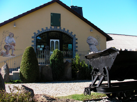 Gelbes Gebäude mit Wandmalereien, grüner Tür und Büschen im Vordergrund; rechts ein schwarzer Kohlewagen auf Schienen.