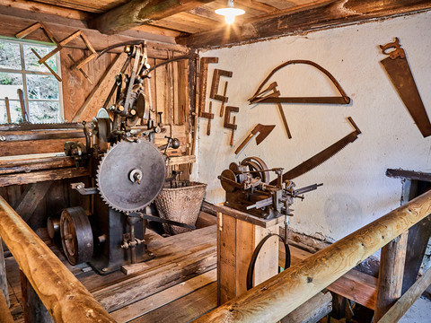 Austellung Neumannmühle Alte Werkstatt mit Holzsägemaschine, umgeben von hölzernen Wänden und Werkzeugen an der Wand; rustikale, historische Atmosphäre.Old workshop with wood sawing machine, surrounded by wooden walls and tools on the wall; rustic, historical atmosphere.Stará dílna s pilou na dřevo, obklopená dřevěnými stěnami a nástroji na stěnách; rustikální, historická atmosféra.Stary warsztat z maszyną do cięcia drewna, otoczony drewnianymi ścianami i narzędziami na ścianie; rustykalna, historyczna atmosfera.Oude werkplaats met houtzaagmachine, omringd door houten muren en gereedschap aan de muur; rustieke, historische sfeer.Vecchia officina con segatrice per il legno, circondata da pareti in legno e attrezzi a muro; atmosfera rustica e storica.