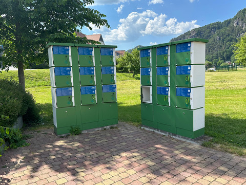 Schließfächer mit Lademöglichkeit Grüne Fahrradschließfächer stehen auf einem gepflasterten Platz im Freien, umgeben von Gras und Bäumen unter einem blauen Himmel mit Wolken.Green bicycle lockers stand on a paved outdoor square, surrounded by grass and trees under a blue sky with clouds.Zelené skříňky na kola stojí na vydlážděném venkovním náměstí, obklopeném trávou a stromy pod modrou oblohou s mraky.Zielone szafki na rowery stoją na wybrukowanym placu, otoczone trawą i drzewami pod błękitnym niebem z chmurami.Groene fietskluisjes staan op een verhard plein in de openlucht, omringd door gras en bomen onder een blauwe lucht met wolken.Gli armadietti verdi per le biciclette si trovano su una piazza pavimentata all'aperto, circondata da erba e alberi sotto un cielo blu con nuvole.