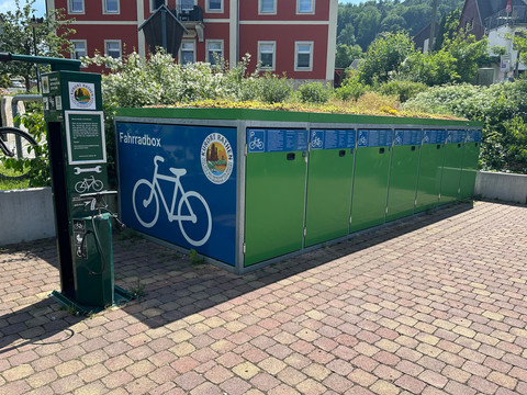 Fahrradboxen in Rathen Grüne Fahrradboxen mit blauer Beschriftung und Fahrradpiktogramm, daneben eine grüne Reparatursäule; im Hintergrund ein rotes Gebäude und grüne Büsche.Green bicycle boxes with blue lettering and bicycle pictogram, next to a green repair column; in the background a red building and green bushes.Zelené boxy na jízdní kola s modrým nápisem a piktogramem jízdního kola vedle zeleného opravárenského sloupu; v pozadí červená budova a zelené keře.Zielone boksy rowerowe z niebieskim napisem i piktogramem roweru, obok zielonej kolumny naprawczej; w tle czerwony budynek i zielone krzewy.Groene fietsdozen met blauwe letters en fietspictogram, naast een groene reparatiezuil; op de achtergrond een rood gebouw en groene struiken.Scatole di biciclette verdi con scritte blu e pittogramma della bicicletta, accanto a una colonna di riparazione verde; sullo sfondo un edificio rosso e cespugli verdi.