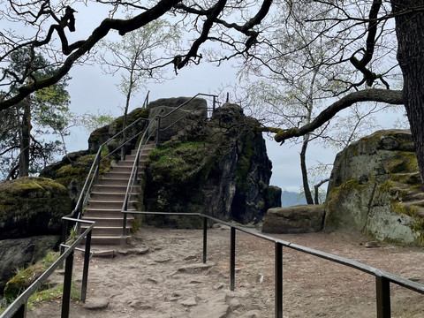 Path to the Canapé View Stone steps with railings lead up to a moss-covered rock, surrounded by bare trees and a gray sky, peaceful atmosphere.
