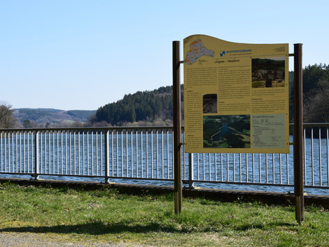 Brucher Talsperre <p>Infotafel vor einem See mit bewaldeten Hügeln im Hintergrund, umzäunt von einem Metallgeländer.</p>