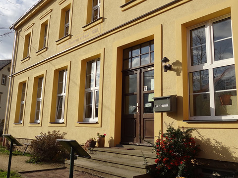 Gelbes Gebäude mit großen Fenstern und einer braunen Tür, daneben ein Briefkasten; sonnige Atmosphäre mit blühenden Sträuchern im Vordergrund.