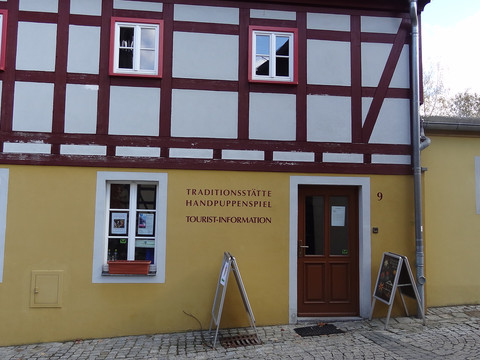 Touristinformation Hohnstein Fachwerkhaus mit gelber Fassade, roten Balken und weißen Fenstern; Aufschrift "Traditionsstätte Handpuppenspiel Tourist-Information" neben Holztür.Half-timbered house with yellow façade, red beams and white windows; inscription "Traditionsstätte Handpuppenspiel Tourist-Information" next to wooden door.Hrázděný dům se žlutou fasádou, červenými trámy a bílými okny; vedle dřevěných dveří nápis "Traditionsstätte Handpuppenspiel Tourist-Information".Dom z muru pruskiego z żółtą fasadą, czerwonymi belkami i białymi oknami; napis "Traditionsstätte Handpuppenspiel Tourist-Information" obok drewnianych drzwi.Vakwerkhuis met gele gevel, rode balken en witte ramen; opschrift "Traditionsstätte Handpuppenspiel Tourist-Information" naast houten deur.Casa a graticcio con facciata gialla, travi rosse e finestre bianche; scritta "Traditionsstätte Handpuppenspiel Tourist-Information" accanto alla porta in legno.
