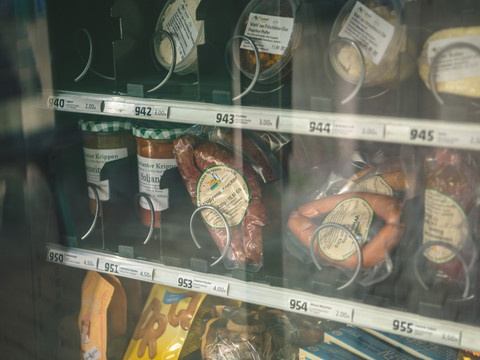 Proviantomat Verkaufsautomat mit verschiedenen Lebensmitteln, darunter Wurstwaren, Suppen im Glas und Snacks, in einem Regal mit Preisschildern.Vending machine with various food products, including sausages, soups in a jar and snacks, on a shelf with price tags.Automat s různými potravinami, včetně párků, polévek ve sklenici a občerstvení, na polici s cenovkami.Automat z różnymi produktami spożywczymi, w tym kiełbaskami, zupami w słoiku i przekąskami, na półce z metkami z cenami.Verkoopautomaat met verschillende voedingsproducten, waaronder worstjes, soepen in een potje en snacks, op een plank met prijskaartjes.Distributore automatico con vari prodotti alimentari, tra cui salsicce, zuppe in barattolo e snack, su uno scaffale con cartellini dei prezzi.
