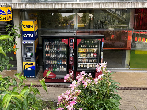 Geldautomat und zwei Snackautomaten vor einem Gebäude mit Glasfassade, umgeben von grünen Pflanzen und rosa Blüten.