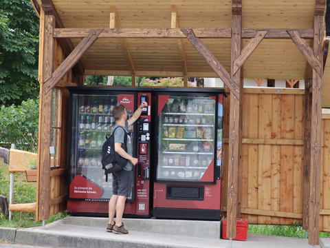 Proviantomat in Pfaffendorf Ein Mann mit Rucksack bedient einen roten Getränke- und Snackautomaten unter einem hölzernen Unterstand.A man with a rucksack operates a red drinks and snack vending machine under a wooden shelter.Muž s batohem obsluhuje červený automat na nápoje a občerstvení pod dřevěným přístřeškem.Mężczyzna z plecakiem obsługuje czerwony automat z napojami i przekąskami pod drewnianą wiatą.Een man met een rugzak bedient een rode drank- en snackautomaat onder een houten afdak.Un uomo con uno zaino gestisce un distributore automatico di bevande e snack rossi sotto una tettoia di legno.