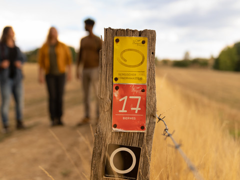 Bierweg Nahaufnahme eines Holzzauns mit Wanderwegschildern; unscharfe Wanderer im Hintergrund auf einem Pfad.
