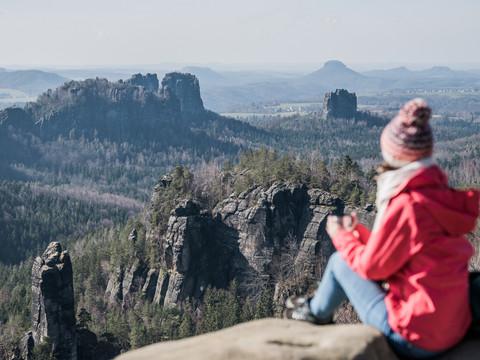 Winterliche Aussicht vom Carolafelsen  Eine Person in roter Jacke und Mütze sitzt auf einem Felsen und blickt auf eine bewaldete Berglandschaft mit markanten Felsformationen.