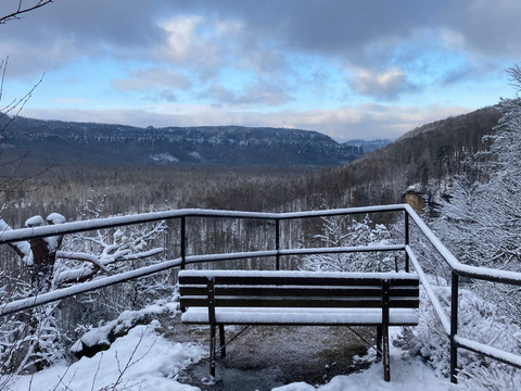Ausblick vom Großstein im Winter Verschneite Bank mit Blick auf ein bewaldetes Tal und entfernte Hügel unter einem bewölkten Himmel mit blauen Lücken.Snow-covered bench overlooking a wooded valley and distant hills under a cloudy sky with blue gaps.Zasněžená lavička s výhledem na zalesněné údolí a vzdálené kopce pod zataženou oblohou s modrými mezerami.Ośnieżona ławka z widokiem na zalesioną dolinę i odległe wzgórza pod zachmurzonym niebem z niebieskimi przerwami.Besneeuwde bank met uitzicht op een beboste vallei en heuvels in de verte onder een bewolkte hemel met blauwe openingen.Panchina innevata con vista su una valle boscosa e colline lontane, sotto un cielo nuvoloso con squarci blu.