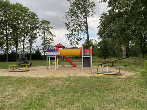 Playground at the Rathewalde adventure pool Spielplatz mit roter Rutsche, gelbem Tunnel, Wippe und Karussell auf Sandfläche, umgeben von Bäumen und Wiese unter bewölktem Himmel.Playground with red slide, yellow tunnel, seesaw and merry-go-round on a sandy surface, surrounded by trees and meadow under a cloudy sky.Dětské hřiště s červenou skluzavkou, žlutým tunelem, houpačkou a kolotočem na písečném povrchu, obklopené stromy a loukou pod zamračenou oblohou.Plac zabaw z czerwoną zjeżdżalnią, żółtym tunelem, huśtawką i karuzelą na piaszczystej powierzchni, otoczony drzewami i łąką pod zachmurzonym niebem.Speeltuin met rode glijbaan, gele tunnel, wip en draaimolen op een zandvlakte, omringd door bomen en weiland onder een bewolkte hemel.Parco giochi con scivolo rosso, tunnel giallo, altalena e girotondo su una superficie sabbiosa, circondata da alberi e prati sotto un cielo nuvoloso.