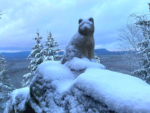 Steinerne Katzenstatue auf schneebedecktem Felsen, umgeben von verschneiten Bäumen und einem weiten, bewaldeten Tal im Hintergrund.