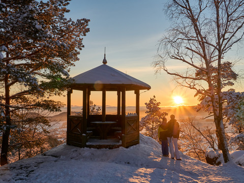 Ein Pavillon im verschneiten Wald bei Sonnenuntergang, zwei Personen stehen nebeneinander und blicken auf die untergehende Sonne.A pavilion in a snowy forest at sunset, two people standing next to each other and looking at the setting sun.Pavilon v zasněženém lese při západu slunce, dva lidé stojí vedle sebe a dívají se na zapadající slunce.Pawilon w zaśnieżonym lesie o zachodzie słońca, dwoje ludzi stojących obok siebie i patrzących na zachodzące słońce.Een paviljoen in een besneeuwd bos bij zonsondergang, twee mensen staan naast elkaar en kijken naar de ondergaande zon.Un padiglione in una foresta innevata al tramonto, due persone in piedi l'una accanto all'altra che guardano il sole che tramonta.