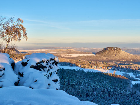 Verschneite Felsen und ein kahler Baum im Vordergrund, mit Blick auf eine winterliche, sonnenbeschienene Hügellandschaft unter klarem, blauem Himmel.