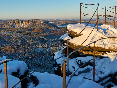 Winter auf dem Gohrisch Verschneite Felsformation mit Geländer im Vordergrund, dahinter ein bewaldetes Tal und ein sonniger, klarer Himmel.