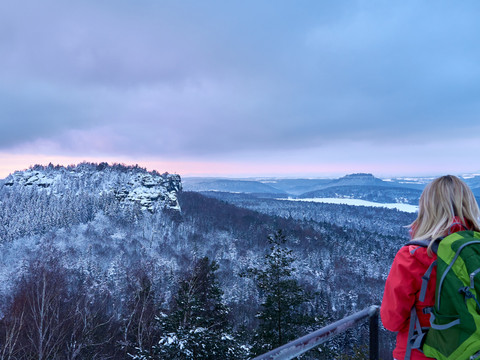 Eine Person mit rotem Rucksack blickt von einem Aussichtspunkt auf eine verschneite Berglandschaft.