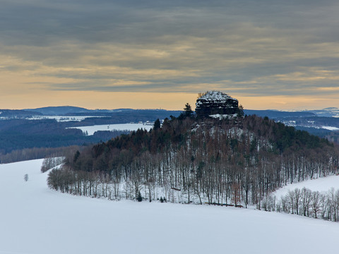 Blick von der Kaiserkrone zum Zirkelstein Schneebedeckte Hügel und karge Bäume vor einer felsigen Klippe, umgeben von weiten Ebenen im Winter.