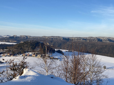 Blick vom Zirkelstein Verschneite Landschaft mit Hügeln im Hintergrund, blauer Himmel, im Vordergrund Bäume und ein Dorf mit bunten Häusern.