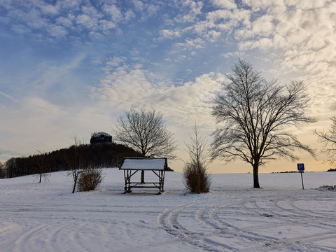 Verschneite Landschaft mit einem hölzernen Unterstand, kahlen Bäumen und einem Parkplatzschild, unter einem bewölkten Himmel bei Sonnenuntergang.