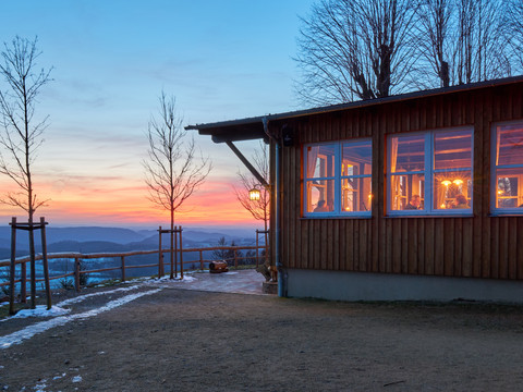 Wachberg-Baude im Winter Holzhaus mit erleuchteten Fenstern bei Sonnenuntergang, umgeben von kahlen Bäumen und einer verschneiten Landschaft.