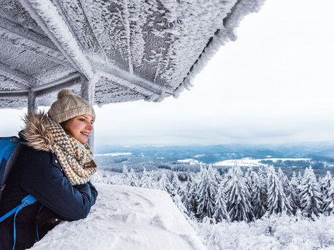 Aussicht genießen auf dem Tanzplan Frau in Winterkleidung mit Mütze und Schal lehnt an einem schneebedeckten Geländer, blickt lächelnd auf verschneite Landschaft und Wälder.