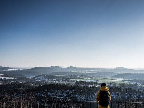 Person in gelber Jacke mit Rucksack blickt von einem Aussichtspunkt auf eine winterliche, hügelige Landschaft unter klarem, blauem Himmel.