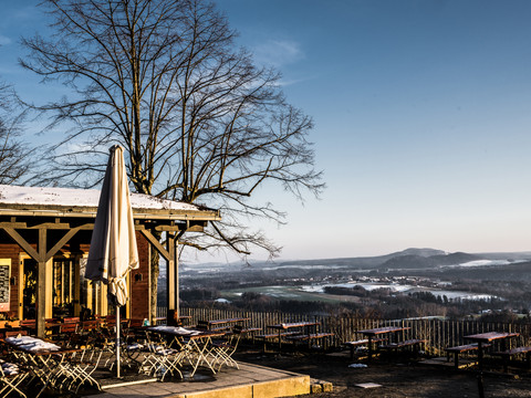 Brandbaude Terrasse eines Cafés mit leeren Tischen und Stühlen, schneebedecktem Dach und Sonnenschirm, im Hintergrund eine winterliche Landschaft unter blauem Himmel.