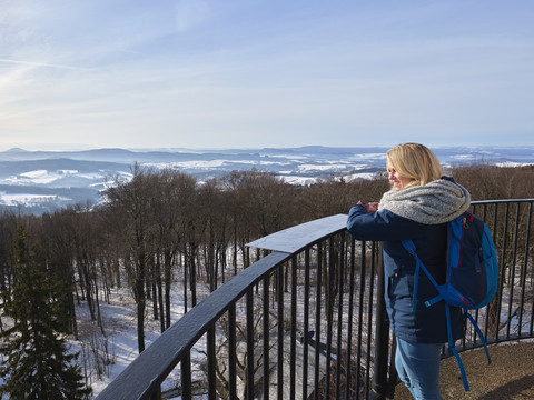 Aussicht vom Ungerberg Eine Frau mit blauem Rucksack steht auf einem Aussichtspunkt und blickt auf eine verschneite Landschaft mit Wäldern und Hügeln.