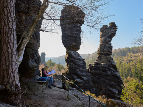 Eine Person sitzt auf einer Bank und liest eine Karte, umgeben von den imposanten Felsformationen der Herkulessäulen und einem bewaldeten Tal unter klarem Himmel.