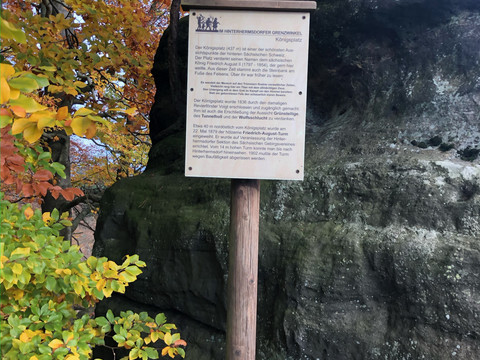 Nápis na náměstí Königsplatz u Hinterhermsdorfu Holzschild mit Text vor einem großen Felsen, umgeben von herbstlich gefärbten Bäumen. Die Atmosphäre ist ruhig und natürlich.