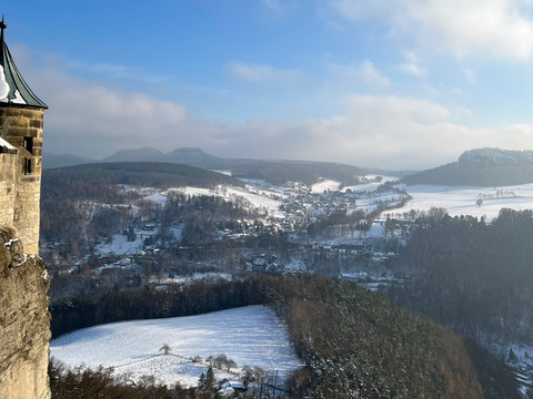Ausblick von der Festung Königstein Verschneite Landschaft mit einem Turm der Festung Königstein links im Vordergrund, sanfte Hügel und ein Dorf im Tal unter blauem Himmel.