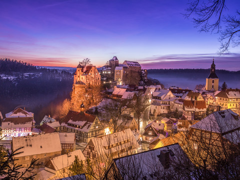 Snow-covered town at sunset with illuminated half-timbered houses, a church and a castle on a hill surrounded by forest.