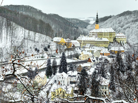 Verschneite Stadtlandschaft mit pastellfarbenen Häusern und einer gelben Kirche auf einem Hügel, umgeben von schneebedeckten Wäldern.